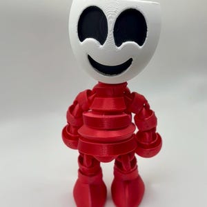 Puede incluir: Una figura articulada impresa en 3D con una cabeza blanca que presenta grandes cuencas oculares negras y una boca ancha sonriente. El cuerpo, los brazos y las piernas están construidos con piezas de plástico rojo segmentadas. El texto "Full color option shown here" es visible.