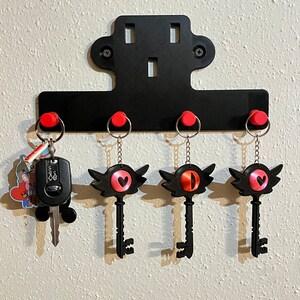 May include: Black key-shaped keychains with red heart and eye designs hang on a black metal key rack with red hooks. The key rack is mounted on a white wall.