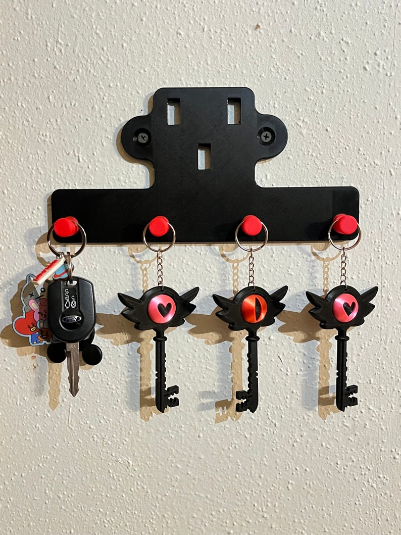 May include: Black key-shaped keychains with red heart and eye designs hang on a black metal key rack with red hooks. The key rack is mounted on a white wall.