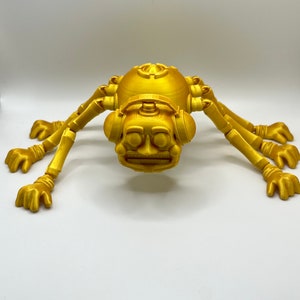 以下が含まれることがあります： 丸い体と8本の脚を持つ、金色の3Dプリントされた関節式クモ。クモは大きな目と広い口を持つ顔をしています。クモはヘッドフォンをしています。