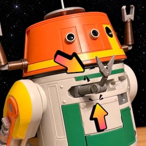 May include: A plastic robot figure featuring an orange dome head with two circular eyes, a white cylindrical body with green panels, and yellow shoulder accents. A small grey mechanical arm extends from the centre, highlighted by two colourful arrows.