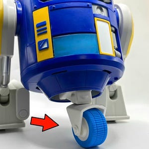 May include: A blue and white robotic toy figure featuring yellow rectangular accents, a central blue translucent panel, and a white wheel assembly at the base. A red arrow points toward the blue wheel, highlighting the mobility component.