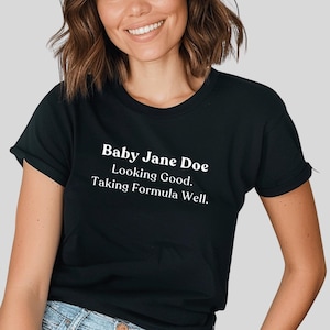 Puede incluir: Camiseta negra con el texto "Baby Jane Doe Looking Good. Taking Formula Well." El texto está en blanco, con una fuente sans-serif. La camiseta tiene mangas cortas y cuello redondo.
