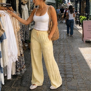 May include: A white spaghetti strap tank top paired with loose-fitting pale yellow trousers featuring an elastic waistband and side pockets. The outfit is accessorized with a woven tan shoulder bag and white closed-toe flat shoes with a textured pattern.