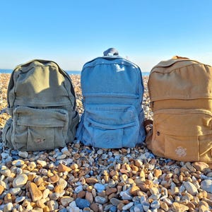 May include: Three canvas backpacks in different colours: green, blue and brown. Each backpack has a front pocket and a small embroidered turtle logo.