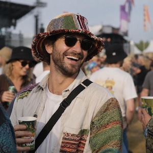 May include: A bucket hat featuring a multi-colored patchwork design with frayed edges, worn with a light-colored button-down shirt that has patterned fabric panels on the shoulders and sleeves, paired with dark-rimmed sunglasses.