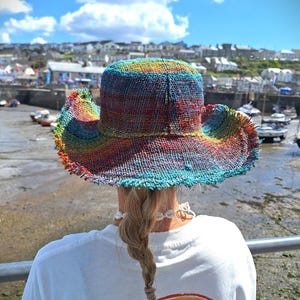 Könnte beinhalten: Ein farbenfroher gewebter Sonnenhut mit breiter Krempe und ausgefransten Kanten. Der Hut besteht aus verschiedenen Farben, darunter Rot, Orange, Gelb, Grün, Blau und Lila.
