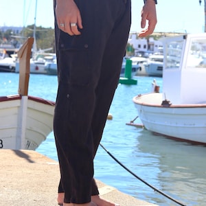 May include: Dark brown cargo trousers with a drawstring waist and side pockets. The trousers are worn barefoot on a concrete dock with boats and water in the background.
