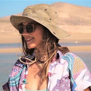 Puede incluir: Una mujer con un sombrero marrón de ala ancha con un logotipo dorado, gafas de sol y una camisa de estampado colorido. Está de pie frente a una playa de arena.