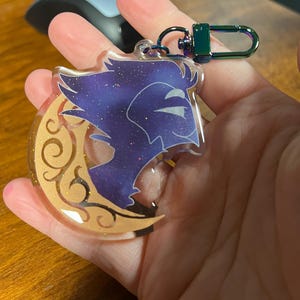 May include: A purple and gold glitter keychain featuring a crescent moon with a stylized wolf silhouette. The keychain has a rainbow iridescent metal clasp.