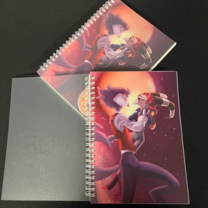 May include: Two spiral bound notebooks with a purple and red character illustration on the cover. The illustration features two characters with horns and tails, one in a red suit and the other in a black suit. The background is a dark purple with a red moon and stars.