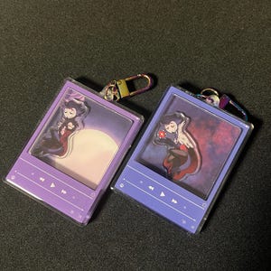 May include: Two acrylic keychains featuring anime-style characters. The keychains have a purple and blue color scheme, with a rectangular design resembling a music player. Each keychain has a different background and character design.
