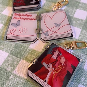 May include: Several acrylic book-shaped keychains with gold-colored clasps. One keychain has the text "Body in abyss Heart in paradise". Other keychains feature illustrations of butterflies, flowers, and two figures in red robes.