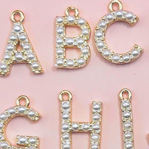 Pu&ograve; includere: Primo piano di ciondoli a forma di lettera in metallo dorato, ciascuno incrostato con piccole perle finte bianche. Le lettere A, B, C, G, H e I sono visibili su uno sfondo rosa tenue. Ogni ciondolo ha un piccolo anello nella parte superiore per il fissaggio.