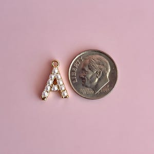 Pu&ograve; includere: Un ciondolo a forma di lettera "A" color oro, ornato di perle finte bianche, accanto a una moneta da 10 centesimi americana. Il ciondolo ha un piccolo anello nella parte superiore per l'attacco. La moneta &egrave; del 2007.