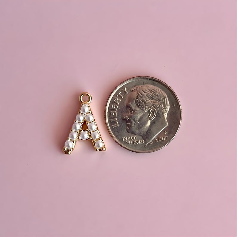 Pu&ograve; includere: Un ciondolo a forma di lettera "A" color oro, ornato di perle finte bianche, accanto a una moneta da 10 centesimi americana. Il ciondolo ha un piccolo anello nella parte superiore per l'attacco. La moneta &egrave; del 2007.