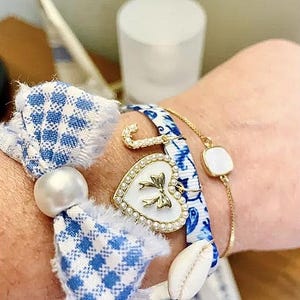 Pu&ograve; includere: Braccialetti con nastro a fantasia blu e bianco, catena dorata e ciondolo a cuore bianco con fiocco dorato. Un braccialetto ha un accento di perla e un ciondolo a conchiglia. I braccialetti sono adornati con dettagli dorati e perle.