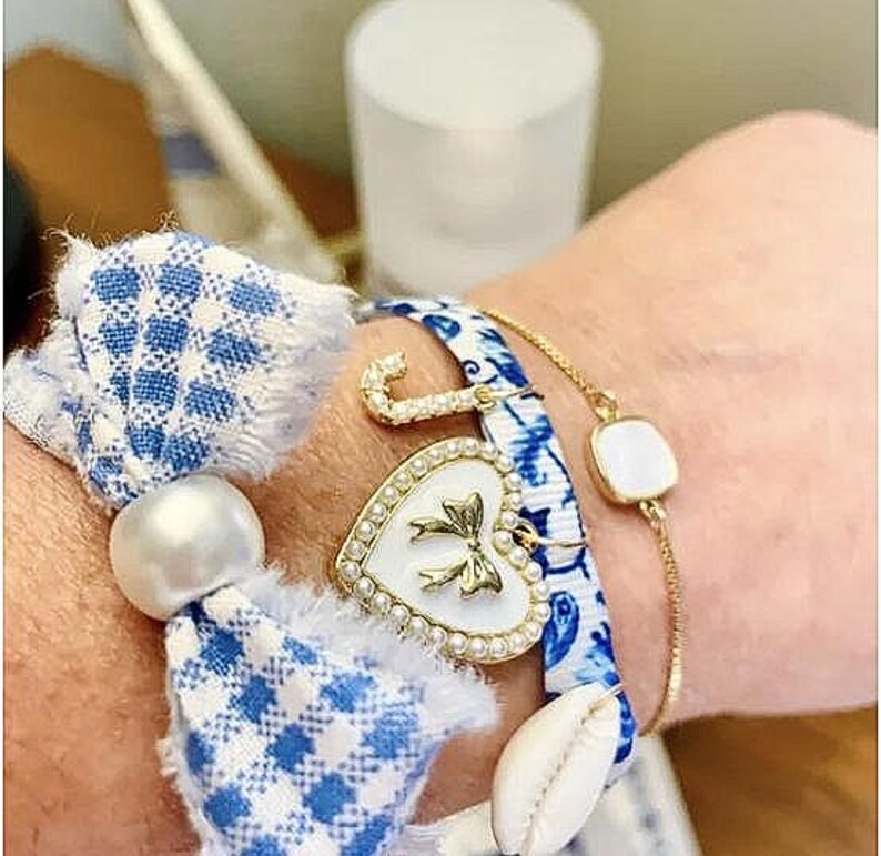 Pu&ograve; includere: Braccialetti con nastro a fantasia blu e bianco, catena dorata e ciondolo a cuore bianco con fiocco dorato. Un braccialetto ha un accento di perla e un ciondolo a conchiglia. I braccialetti sono adornati con dettagli dorati e perle.