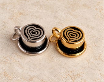 Puede incluir: Dos dijes de metal con forma de tazas de café sobre platillos. Un dije es de tono plateado con un diseño de arte latte en forma de corazón negro, y el otro es de tono dorado con un diseño de arte latte en forma de corazón negro. Incluye argolla.