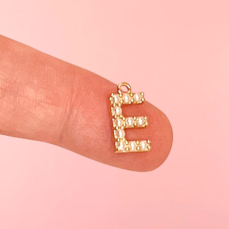 Pu&ograve; includere: Un ciondolo dorato a forma di lettera "E", incastonato con piccole pietre preziose trasparenti. Il ciondolo ha un piccolo anello nella parte superiore per attaccarlo a una collana o un braccialetto. Il ciondolo &egrave; mostrato su uno sfondo rosa.