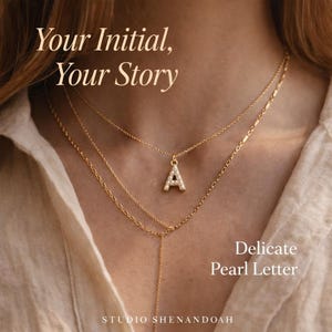 Pu&ograve; includere: Collane dorate a strati con un ciondolo iniziale tempestato di perle. La lettera "A" &egrave; il punto focale, con il testo "Your Initial, Your Story" e "Delicate Pearl Letter". Il logo Studio Shenandoah &egrave; in basso.