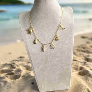Puede incluir: Collar dorado con varios colgantes tem&aacute;ticos del oc&eacute;ano, incluyendo una tortuga, una concha, un delf&iacute;n y una br&uacute;jula. El collar se exhibe sobre un busto blanco contra un fondo de playa, con arena y mar visibles.
