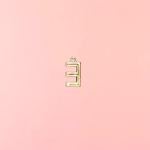 Pu&ograve; includere: Un ciondolo in metallo color oro a forma di lettera "E" su sfondo rosa tenue. Il ciondolo ha un piccolo anello nella parte superiore per il fissaggio a una collana o un braccialetto. La lettera &egrave; in un font sans-serif.