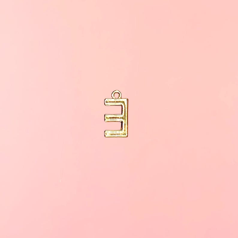 Pu&ograve; includere: Un ciondolo in metallo color oro a forma di lettera "E" su sfondo rosa tenue. Il ciondolo ha un piccolo anello nella parte superiore per il fissaggio a una collana o un braccialetto. La lettera &egrave; in un font sans-serif.