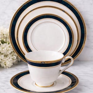 Puede incluir: Elegante juego de té de porcelana blanca con un diseño de borde azul marino y dorado. El juego incluye una taza de té, un platillo y platos de varios tamaños. El juego de té se exhibe sobre una superficie de mármol.