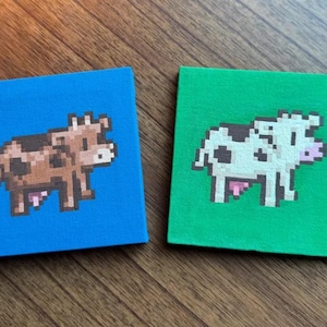 May include: Two square canvases with pixelated cow illustrations. One canvas is blue with a brown cow, the other is green with a black and white cow. The artwork is reminiscent of retro video game graphics.