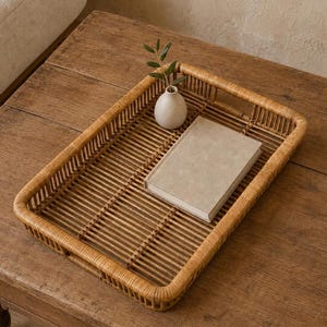 Puede incluir: Una bandeja rectangular de ratán trenzado con acabado marrón claro. Dentro de la bandeja hay un pequeño jarrón de cerámica color blanco roto con una rama de hojas verdes, junto a un libro rectangular cerrado de tapa beige.