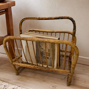 May include: A weathered, light brown bamboo magazine rack with a curved handle and vertical slats. The structure features a distressed finish with darker brown patches throughout. Several books with neutral-toned covers are stored inside the rack.