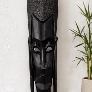 May include: A tall, black wooden mask with a stylized face. The mask has a long, rectangular shape with a textured top and a prominent nose and mouth. The mask is displayed against a white wall with a potted plant.