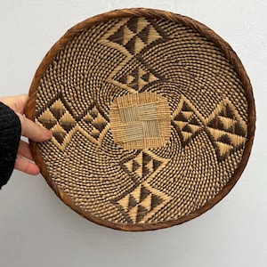 May include: Round woven basket with a brown rim and intricate geometric patterns in shades of brown and beige. The basket's design features diamond and triangle shapes, creating a textured surface.