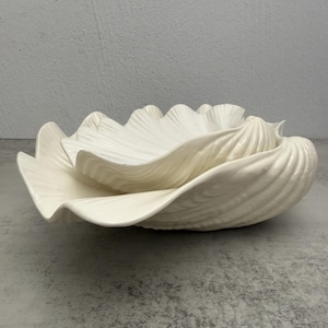 May include: A cream-colored ceramic bowl shaped like a large, open seashell. The bowl features a glossy finish with deep, radiating ridges that mimic the natural texture of a clam shell, creating a wide, scalloped edge and a deep, curved basin.