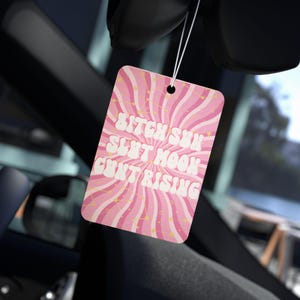 May include: Pink air freshener with the text "Bitch Sun Slut Moon Cunt Rising" in white. The air freshener has a retro design with pink and white wavy lines and starbursts. It is hanging in a car.