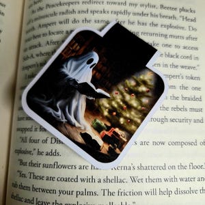 May include: A white ghost-shaped bookmark with a spooky illustration. The image shows a ghost holding a book with a Christmas tree and presents in the background. The bookmark is placed on top of a book.