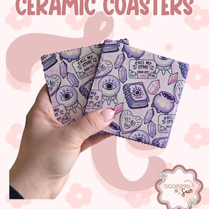 May include: Two ceramic coasters with a light purple background and a repeating pattern of crystal balls, books, and other mystical symbols. The coasters have the text "CERAMIC COASTERS" at the top.