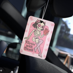 May include: A pink air freshener with a cartoon skeleton illustration. The skeleton is wearing heart-shaped sunglasses and holding a heart. The text "Flirtin' with Death" is printed on the bottom.