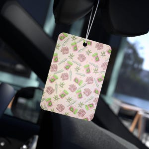 May include: Rectangular air freshener with a repeating pattern of pink and green cannabis-themed illustrations. The design includes jars, buds, and leaves on a light pink background. The air freshener is hanging in a car.
