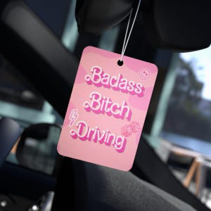May include: A pink car air freshener with the words "Badass Bitch Driving" in white and pink lettering. The air freshener has a camouflage pattern and a small smiley face and dice graphic.