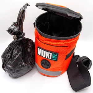 Può includere: Dispenser per sacchetti per la raccolta delle feci dei cani di colore arancione con una cinghia nera e un coperchio nero. Il dispenser ha una striscia riflettente e un logo che dice "Muki Life".