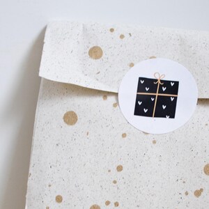 Op de afbeelding: Een gevouwen papieren cadeauzakje met een gespikkelde, gebroken witte achtergrond en gouden verfspatten. Een ronde sticker met een zwart-wit cadeau-ontwerp is op de voorkant bevestigd. De tas rust op een licht houten oppervlak.