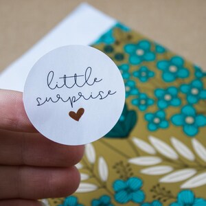 Op de afbeelding: Een witte ronde sticker met de woorden "little surprise" in zwart script en een klein bruin hart. De sticker wordt boven een achtergrond met patronen met turquoise bloemen en een goudkleurige basis gehouden.
