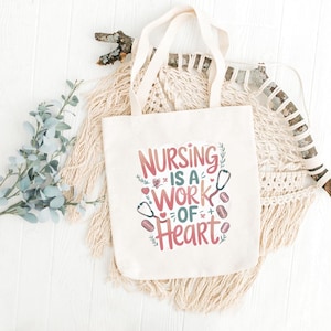 Op de afbeelding: Crèmekleurige draagtas met de tekst "NURSING IS A WORK OF HEART" in roze, rood en groen. Het ontwerp bevat stethoscopen en harten. De tas heeft crèmekleurige handvatten en wordt gepresenteerd op een witte ondergrond.