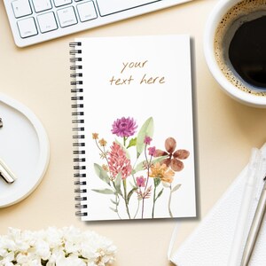 Pode incluir: Um caderno espiralado branco com uma capa com um design floral em aquarela em tons de rosa, roxo e laranja. O texto "your text here" está escrito em script marrom. Uma xícara de café e um teclado estão no fundo.