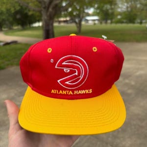 May include: A red baseball cap with a yellow brim featuring the white embroidered Atlanta Hawks logo and the text Atlanta Hawks in yellow. The cap includes yellow eyelets on the crown and a structured front panel with a classic snapback design.