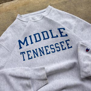 May include: A light gray crewneck sweatshirt featuring the text MIDDLE TENNESSEE in bold blue block letters across the chest. The garment has a ribbed collar, cuffs, and hem, with a small embroidered Champion logo patch on the left sleeve cuff.