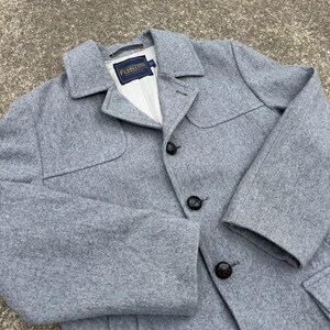 Puede incluir: Un abrigo de mezcla de lana gris claro con cuello clásico, tres botones de color marrón oscuro en la parte delantera y una etiqueta de la marca Pendleton cosida en el interior del cuello. La prenda incluye un forro blanco y costuras estructuradas en los hombros.