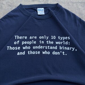 Peut inclure: Un t-shirt bleu marine à manches courtes avec une impression graphique blanche qui dit : There are only 10 types of people in the world: Those who understand binary, and those who don't. Le t-shirt porte une étiquette de col Gildan Ultra Cotton.
